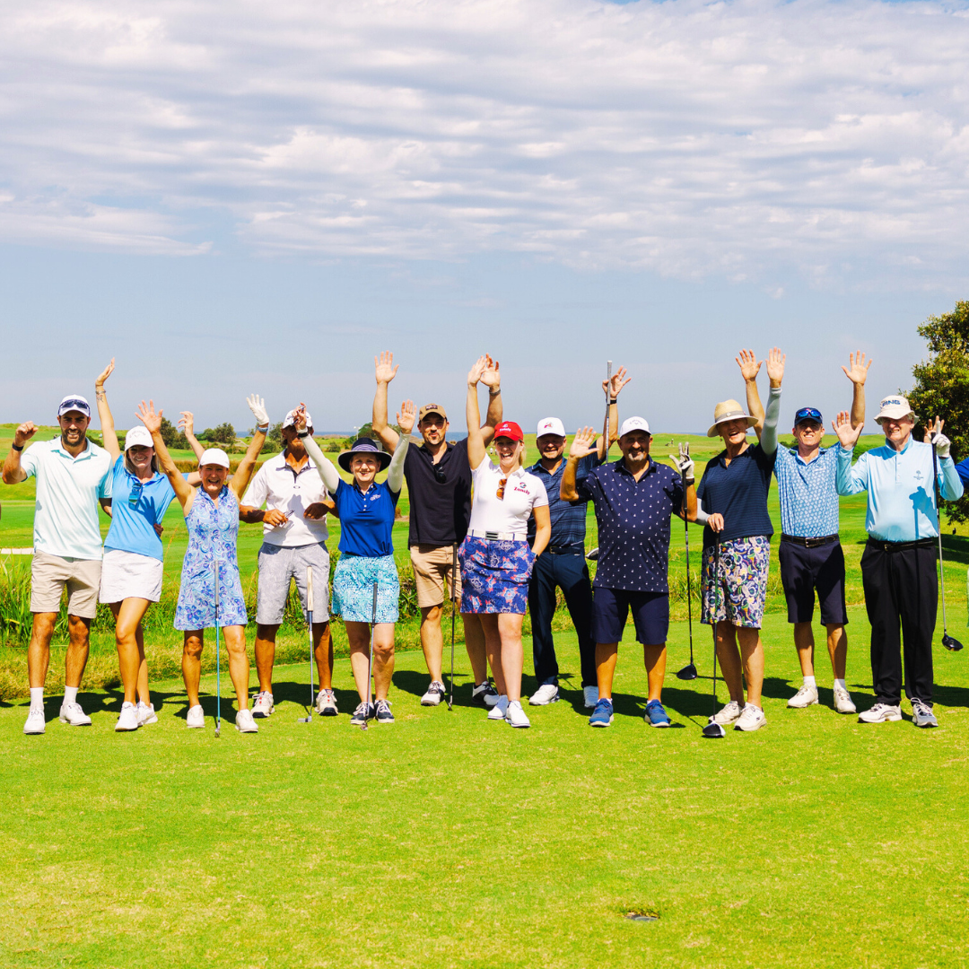 A group of golfers stand together on a sunny course, smiling with arms raised and clubs in hand.